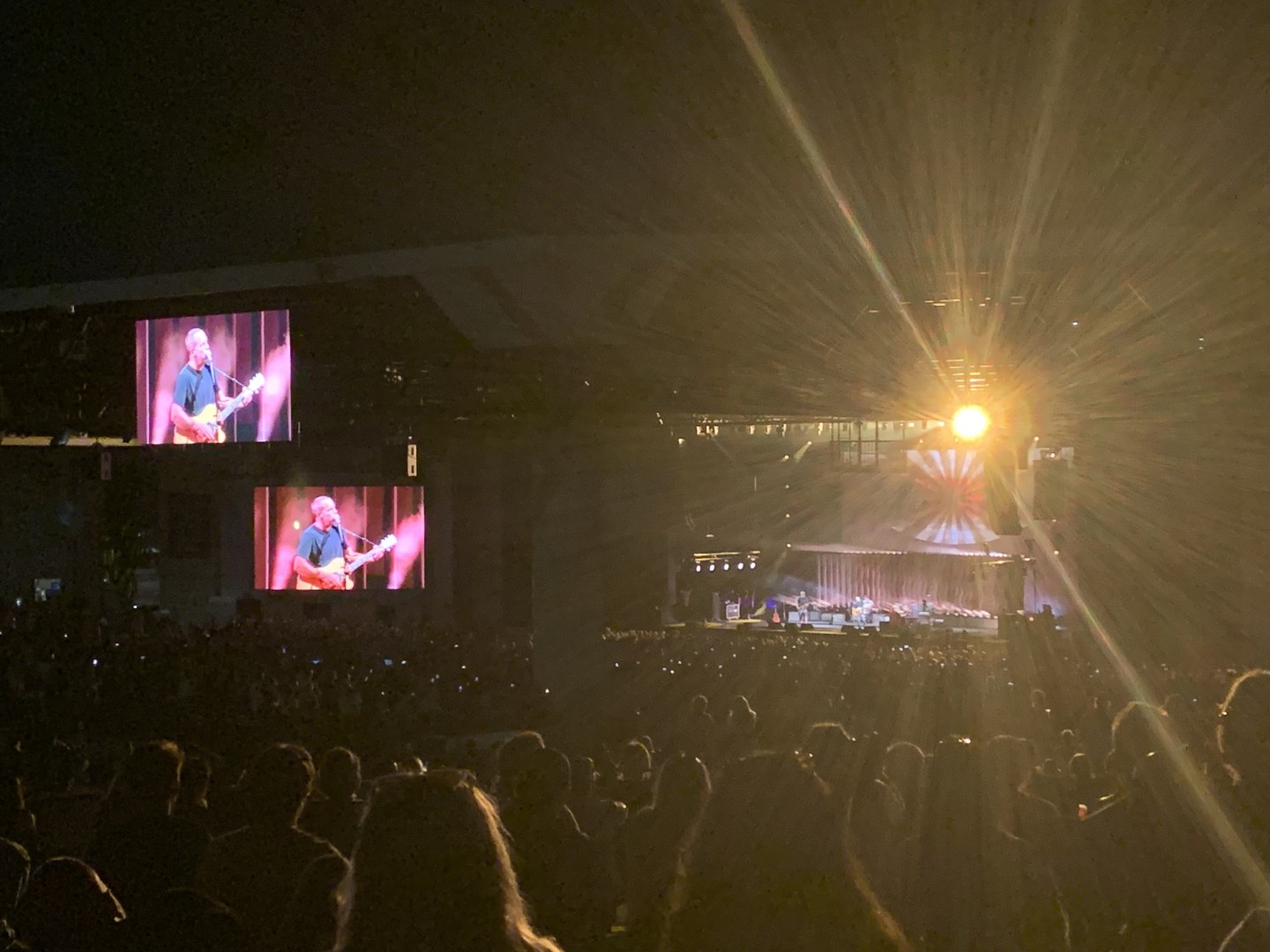 A dimly lit photo of a concert stage and two TVs displaying the stage at an outdoor concert. Jack Johnson is wearing a blue T-shirt and is playing the guitar and is displayed on both Tv screens.